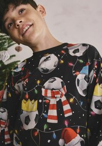 Băiat zâmbind lângă un brad de Crăciun decorat, purtând un pulover negru cu mingi de fotbal și îmbrăcat în pălării de Moș Crăciun, eșarfe, coroane și lumini de sărbătoare.