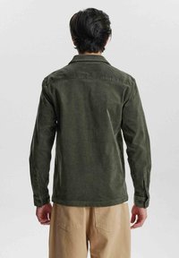 Cămașă din bumbac cord cu mânecă lungă, de culoare verde măsliniu, cu guler, manșete cu button și o croială lejeră, combinată cu pantaloni bej. Se observă textura materialului.