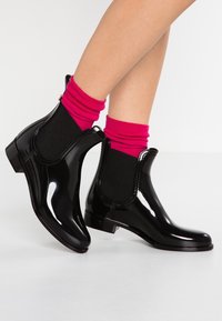 Bottes de pluie noires brillantes à la cheville avec des panneaux élastiques sur les côtés, des petits talons carrés et des coutures décoratives. Chaussettes froncées roses et noires visibles.