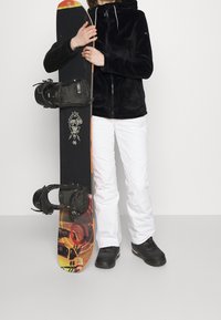 Black fleece jacket with drawstrings, white snow pants, and black snow boots. Holding a snowboard featuring colorful graphics and bindings.