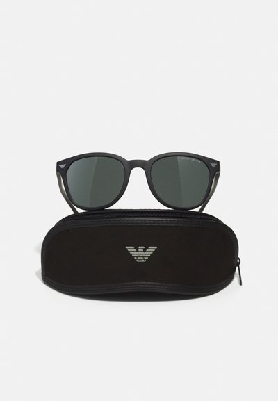 Ochelari de soare negri cu lentile verzi expuși deasupra unui toc negru cu fermoar, având un logo argintiu. Ramele sunt elegante, cu un finisaj mat.