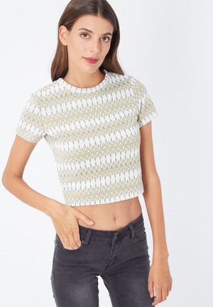 Camiseta corta en blanco con un patrón zigzag amarillo; mangas cortas, cuello redondo, textura acanalada; combinada con vaqueros negros.