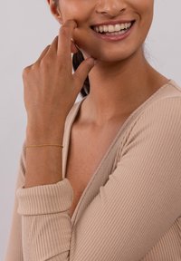 Goldener, gedrehter Armband, der am Handgelenk getragen wird, kombiniert mit einem beigen, gerippten Langarmoberteil mit tiefem V-Ausschnitt. Weiche Haut und strahlendes Lächeln sichtbar.