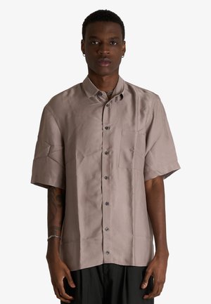 Uomo che indossa una camicia marrone chiaro a maniche corte con taschino sul petto, pantaloni neri, bracciale a catena e orecchini su sfondo bianco.