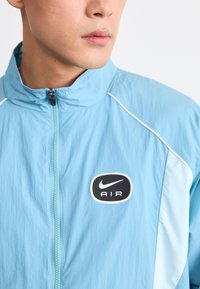 Ljusblå Nike-jacka med halv dragkedja, med svart och vit "AIR"-logotyp på bröstet, minimalistisk design och textil med struktur.
