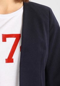 Vit skjorta med stort rött nummer 7 delvis täckt av en marinblå, texturerad kavaj på en person.