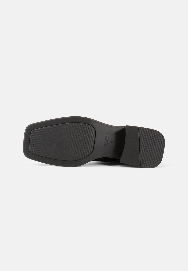 Semelle de chaussure noire avec une surface en caoutchouc texturé, de forme rectangulaire, talon légèrement surélevé et marquage visible sur le côté intérieur.