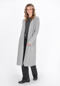 Donna che indossa un lungo cardigan grigio a maglia sopra un top nero e jeans, in piedi con un piede in avanti contro uno sfondo bianco semplice.