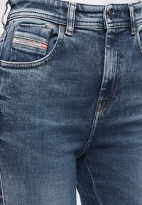 Κοντινό πλάνο σε μπλε τζιν με μπροστινό κουμπί, φερμουάρ, θηλιές για ζώνη και μια μικρή ετικέτα στην τσέπη που γράφει «Diesel Industry Denim Division».