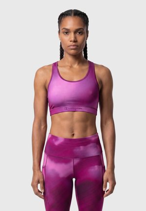 Mujer atlética con trenzas vistiendo un sujetador deportivo morado a juego y leggings de cintura alta sobre un fondo claro liso.