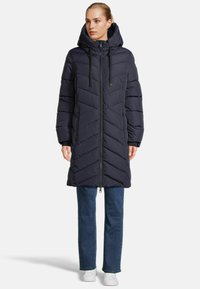 Marineblauer gepolsterter Mantel mit Kapuze, ausgestattet mit einem Reißverschluss vorne, Chevron-Steppung und zwei Seitentaschen. Getragen über Jeans und weißen Schuhen.