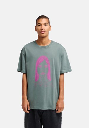 Jonge man met kort haar die een loszittend groen T-shirt draagt met een grote roze lijntekening van een vrouwengezicht en tekst aan de voorkant.