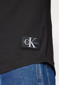 Black cotton t-shirt featuring a white Calvin Klein Jeans logo patch on the bottom left, paired with blue denim jeans visible in the image.