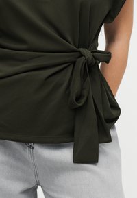 Σκούρο πράσινο αμάνικο τοπ με λεπτομέρεια στη σκέπη στο πλάι, συνδυασμένο με ανοιχτό γκρι τζιν σε άτομο μπροστά σε απλό φόντο.