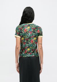 Άτομο με μακριά σκούρα μαλλιά, που φοράει κοντομάνικο μαύρο πουκάμισο με κίτρινα μανίκια και μακριά μαύρη φούστα, στραμμένο με την πλάτη προς τον θεατή.