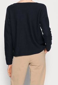 Pull en maille bleu marine avec un large col bateau, des manches longues et une coupe décontractée, associé à un pantalon beige. Texture lisse, design simple.