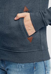 Sudadera gris oscuro con un bolsillo frontal y un acento triangular de cuero marrón. Textura suave, puños ajustados y diseño casual.