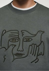 Abstrakcyjna grafika liniowa przedstawiająca twarz z dwiema rękami i dwoma ptakami wkomponowana w projekt na szarej bluzie.