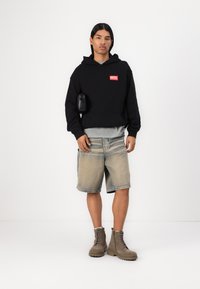 Sort hætte sweatshirt med rødt "DIESEL" logo, lagdelt over en grå t-shirt. Båret med lyse denim shorts og beige snørebåndsstøvler.