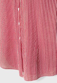 Chemise rayée rouge et blanche avec des lignes verticales, dotée d'une patte de boutonnage au centre et d'un ourlet incurvé. Fabriquée en tissu léger.