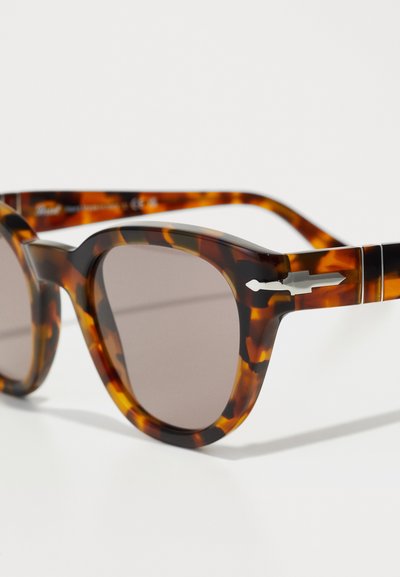 Ochelari de soare din material tartan cu rame rotunde, lentile cu nuanță maro și accente negre elegante pe brațele și curea.