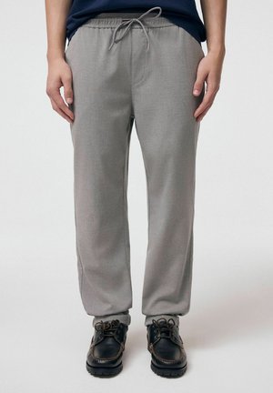 Osoba nosząca jasnoszare spodnie ze sznurkiem i czarne sznurowane buty, stojąca na białym, jednolitym tle.