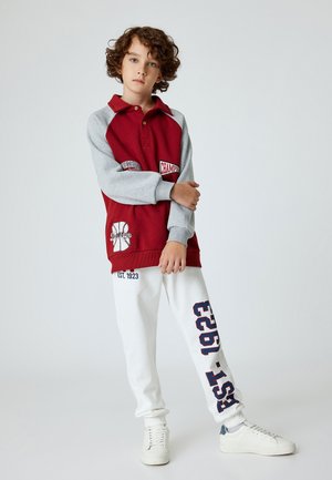 Rød og grå pullover med krave, der har forskellige logoer og broderier. Hvide sweatpants med blå tryk og en afslappet pasform. Hvide sneakers.