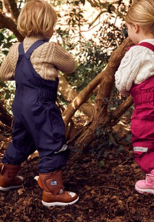 Zwei Kinder stehen zwischen Laub, tragen wasserdichte Overalls; einer in Marineblau mit braunen Stiefeln, der andere in Rosa mit weißen Akzenten.