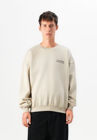 Sweatshirt en coton beige avec un col rond et des bords-côtes. "SOHO Social Coffee CLUB" imprimé en noir sur la poitrine gauche. Texture douce, coupe décontractée.