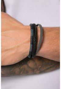 Tre braccialetti su un polso: uno in pelle nera intrecciata, uno in tessuto liscio grigio scuro e uno in tessuto intrecciato nero con accenti blu.