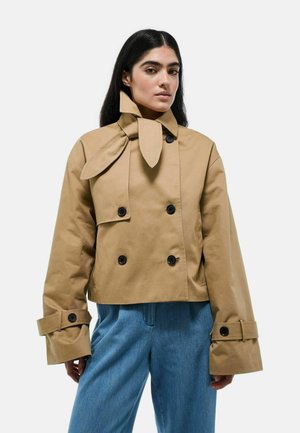 Femme portant une veste croppée beige à double boutonnage avec des poignets surdimensionnés et un col unique noué en nœud, associée à un jean bleu ample.