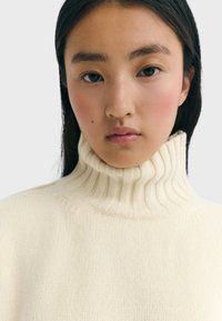 Cremfarbener Rollkragenpullover mit geripptem Muster und umgeschlagenem Kragen, der einen lässigen Schnitt und weiches Strickmaterial aufweist.