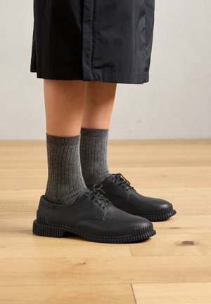 Mustmustest mustad nahkjalatsid tekstuuriga pinnaga ja soontega tallad, koos hallide ribadega sokkidega, puidust põranda taustal.
