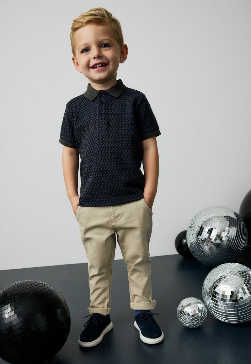 Polo shirt blu navy con un motivo geometrico, pantaloni khaki e scarpe blu navy. Circondato da sfere da discoteca di dimensioni variabili.