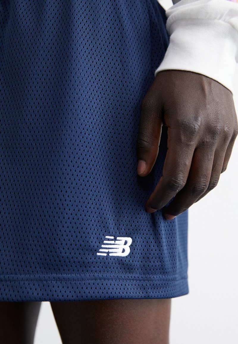Pantalones cortos de malla azul marino con un patrón texturizado y un logo de New Balance bordado en blanco en el lado izquierdo. Mano descansando cerca del dobladillo.
