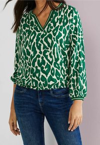 Blusa verde a fantasia con scollo a V, caratterizzata da un audace motivo maculato, con polsini elasticizzati, abbinata a jeans di denim blu.