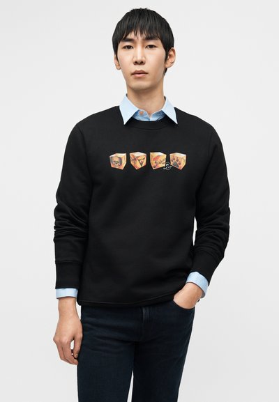 Μαύρο φούτερ με τέσσερα εικονογραφημένα, πολύχρωμα σχέδια κύβων. Φορεμένο πάνω από ανοιχτό μπλε πουκάμισο με εμφανείς μανσέτες. Σκούρο τζιν.