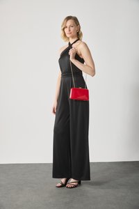 Salopette noire sans manches avec un détail noué au niveau du col, assortie à une pochette rouge brillant et des sandales noires à lanières. Texture lisse.