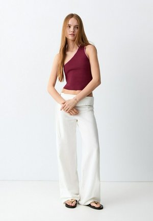 Tânără cu păr lung roșcat, purtând un top bordo fără mâneci, pantaloni albi largi și papuci negri, stând pe un fundal alb simplu.