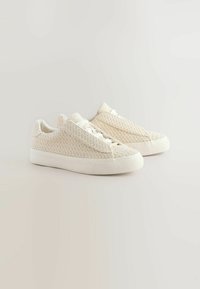 Baskets de couleur crème avec une texture tissée, semelle plate en caoutchouc blanc et fermeture à lacets. Design minimaliste, adapté pour une tenue décontractée.