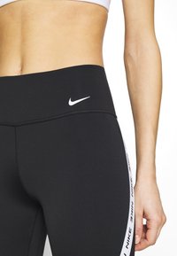 Leggings noirs en tissu extensible, dotés d'une taille haute, du logo Nike à l'avant et d'un ruban logo sur les côtés.