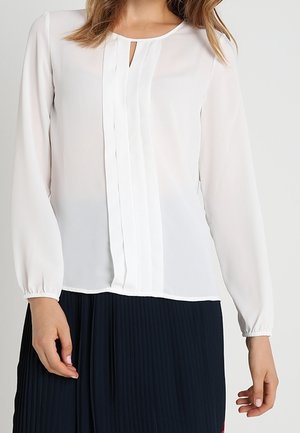 Vrouw draagt een witte blouse met lange mouwen met geplooide voorkant en een keyhole-halslijn, gecombineerd met een donkere geplooide rok.