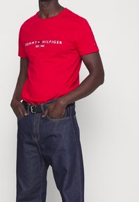 Punainen puuvillapaita, jossa on valkoinen "TOMMY HILFIGER" -logo, käytetään tummansinisten farkkujen ja mustan vyön kera. Lyhyet hihat, pyöreä pääntie.