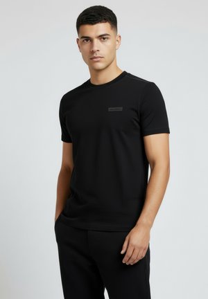 Ung mand med kort mørkt hår iført en ensfarvet sort t-shirt med korte ærmer og sorte bukser, stående mod en ensfarvet lys baggrund.