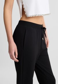 Pantaloni neri con coulisse presentano una comoda vita, una parte anteriore plissettata e dettagli in metallo dorato. Abbinati a un top bianco corto con bordi di strass.