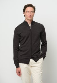 Cárdigan de cremallera gris oscuro con cuello alto, mangas largas y un diseño ajustado. Lleva puesto sobre una blusa de color claro y combinado con unos pantalones claros.