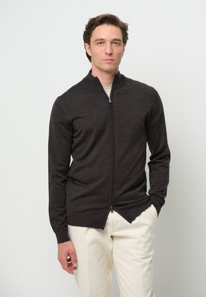 Cárdigan de cremallera gris oscuro con cuello alto, mangas largas y un diseño ajustado. Lleva puesto sobre una blusa de color claro y combinado con unos pantalones claros.