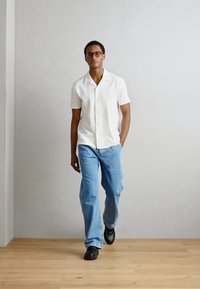 Weißes kurzärmliges Hemd mit einem strukturierten Muster, blaue locker sitzende Jeans und schwarze Schuhe, auf einem neutralen Hintergrund mit einem hellen Holzboden.