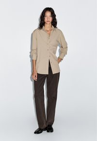 Beige Hemd mit langen Ärmeln, kombiniert mit dunkelbraunen geraden Hosen und schwarzen Schuhen. Glatte Stoffqualität mit einem lockeren Schnitt.