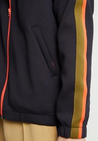 Chaqueta navy con cremallera naranja y dos franjas, verde olivo y naranja, que bajan por la manga, usada sobre pantalones beige.